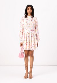 Φορεμα με λουλούδια και μακριά μανίκια, με ριγέ γιακά και εφαρμοστή μέση. Το μοντέλο κρατάει μια ροζ τσάντα. Οι καφέ σανδάλια με λουριά ολοκληρώνουν την εμφάνιση.