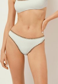 Weiße gerippte Bikiniunterteile mit einer strukturierten Oberfläche und kontrastierendem braunem gewelltem Saum entlang des Taillenbunds und der Beinschnitte.