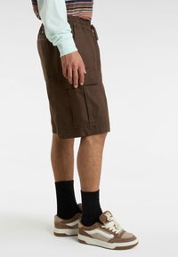 Brązowe szorty cargo z elastycznym pasem, z dwoma dużymi bocznymi kieszeniami; w zestawie z paskowanym, długim rękawem oraz brązowymi sneakersami.