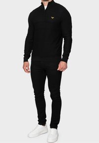 Schwarzer Strickpullover mit einem Viertel-Reißverschluss und strukturiertem Muster. Hat ein kleines gelbes Logo auf der Brust, kombiniert mit schwarzen Skinny-Jeans und weißen Sneakers.