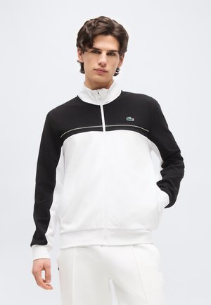 Fiatal férfi fekete-fehér cipzáras dzsekiben, krokodil logóval, egyik kezét a zsebében tartva, egyszerű háttér előtt áll.