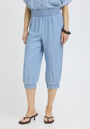 Mujer con pantalones cortos azul claro con cintura elástica y puños, combinados con sandalias negras de tacón y una pulsera rígida.