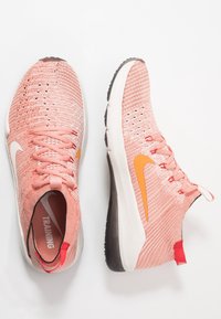 Rosa Nike träningsskor har en texturerad ovandel, orange logotyp och vita sulor, visade från en uppifrån och ner perspektiv på en vit bakgrund.