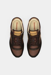 Brune idrettssko med en blanding av materialer på overdelen. Har mørkere brune detaljer, en strukturert svart såle og "Saucony Jazz" merke.