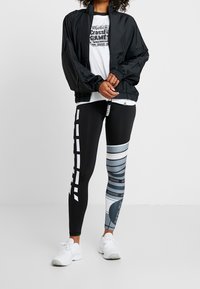 Giacca a vento nera, t-shirt grigia con logo, e leggings a fantasia neri, grigi e blu con dettagli a righe. Scarpe da ginnastica bianche.