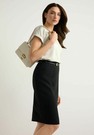 Kobieta ubrana w białą bluzkę i czarną spódnicę, trzymająca beżową torebkę z złotym łańcuszkiem na ramieniu.