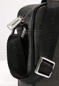 Black synthetic bag with logo pattern, silver hardware, and a woven adjustable strap. Features a top zipper and keychain accents.