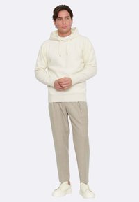 Felpa a maglia di colore crema con cappuccio e cordino, abbinata a pantaloni su misura grigi chiari e sneaker bianche. Design semplice e dal fit rilassato.