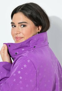 Nainen, jolla on tummat hiukset, pukeutunut tähtikuosiseseen violettiin takkiin, nostamassa kaulusta ylös, hymyilee hieman, yksinkertaisella valkoisella taustalla.