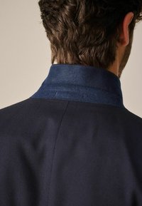 Marineblauwe jas met een gestructureerd lichaam en een contrasterende gladde kraag. Voorzien van stiksels, wat een op maat gemaakte uitstraling benadrukt.