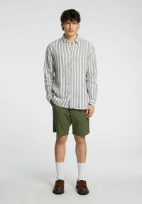 Camicia a maniche lunghe da uomo con righe verticali bianche, blu e beige, abbinata a pantaloni corti color verde oliva. Il modello indossa scarpe marroni e calzini bianchi.