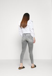 Camisa blanca con cuello rizado, jeans ajustados grises y sandalias negras sin talón. El cabello lacio cae sobre los hombros. Fondo sencillo.