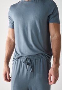 Close-up of a man wearing a gray t-shirt and matching drawstring pants, highlighting fabric texture and fit. Background is neutral.