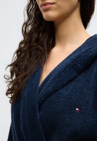 Μπλε ναυτικό χρώμα μπουρνούζι από περκάλι με κουκούλα, με υφή υφάσματος και μικρό λογότυπο σε κόκκινο, λευκό και μπλε στο μπροστινό μέρος.