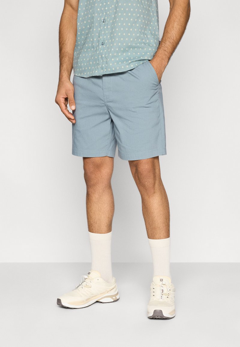 Ανοιχτό μπλε βαμβακερό σορτς με πλαϊνές τσέπες, φορεμένο με πουκάμισο με σχέδια, λευκές κάλτσες και λευκά αθλητικά παπούτσια. Λεία υφή, casual σχεδίαση.