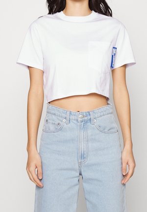 Kobieta w białym krótkim T-shircie z małą kieszonką i niebieską metką, w połączeniu z jasnoniebieskimi jeansami z wysokim stanem.