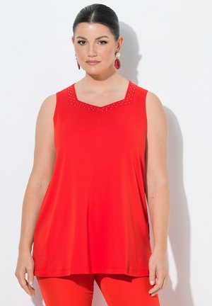 Femme aux cheveux foncés portant un haut rouge sans manches et un pantalon rouge, avec des boucles d’oreilles rouges et en perles, debout contre un mur blanc.