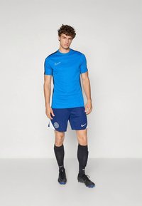 Camiseta deportiva azul de manga corta con un detalle de raya negra, combinada con pantalones cortos azul marino que tienen un logo y un acento blanco. Botas de fútbol negras.