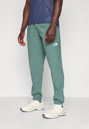Pantalon de jogging léger vert avec poignets élastiques, doté d'un logo blanc sur la cuisse gauche. Accompagné de chaussures de sport beiges.