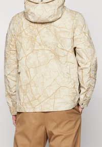 Beige kviltad jacka med en kartmönster i ljusbrunt. Har hög krage, sidofickor och knappade ärmslut. Bärs med beige byxor.