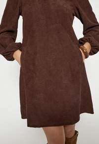 Hnedé, textúrované šaty s voľným strihom, dlhými rukávmi a bočnými vreckami. Materiál má rebrovaný vzor a elastické manžety.