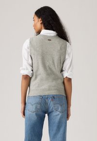 Gilet tricoté gris avec un col rond, en tissu texturé, et une petite étiquette brune à l'arrière, associé à des manches blanches retroussées et un jean bleu.