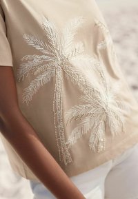 Beige top s krátkým rukávem zdobený bílou výšivkou palmového stromu s perlovými detaily. Hladká textura a volný střih, kombinovaný s bílými kalhotami.