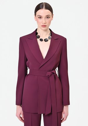 Femme portant un blazer ceinturé violet profond et un pantalon assorti, accessoirisée d’un collier épais en pierre noire, debout devant un fond uni.