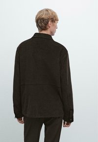 Giacca in corduroy marrone scuro con colletto, cuciture visibili e polsini con bottoni. La parte posteriore presenta un design con cucitura orizzontale.