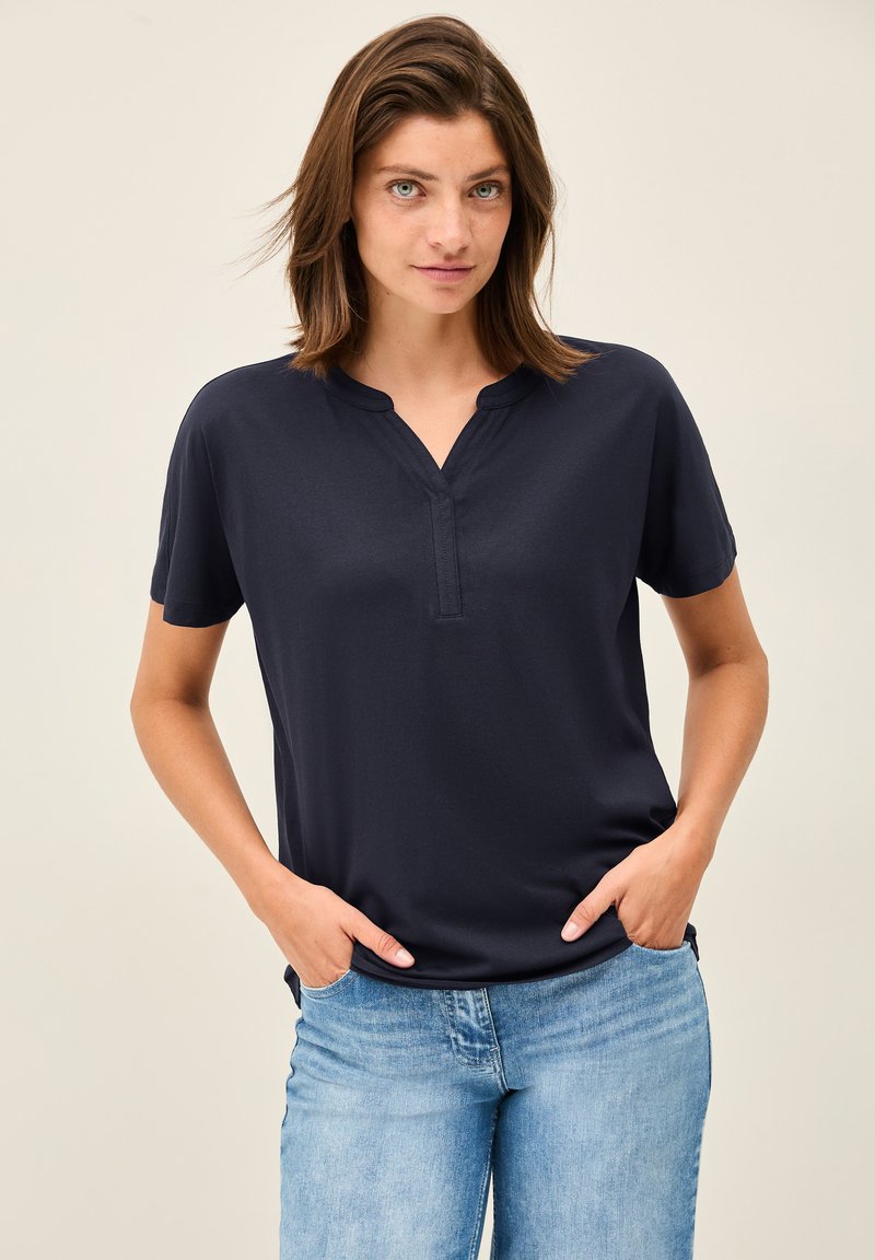 Haut marine à manches courtes avec un col henley ; tissu doux, texture lisse, coupe décontractée, accompagné de jeans en denim bleu clair.