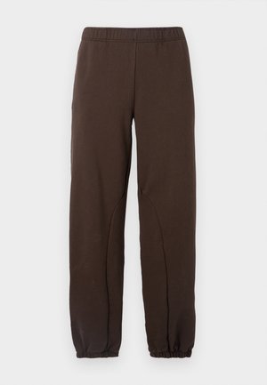 Person trägt braune, locker sitzende Hose und passendes kurzärmliges Hemd über einem schwarzen Langarmshirt, schwarze Schuhe und trägt eine schwarze Umhängetasche.