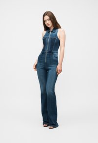 Γυναίκα που στέκεται φορώντας ένα αμάνικο σκούρο μπλε τζιν ολόσωμο φόρεμα με φερμουάρ στο μπροστινό μέρος και φαρδιά μπατζάκια, ποζάροντας μπροστά σε μια απλή φόντο.