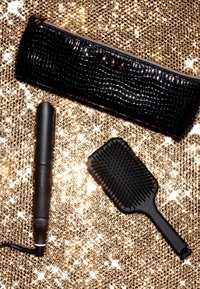 Zwarte krokodillen-embossed pouch met rits, keramische haard straightener in matzwart, en een grote zwarte paddleborstel op een sprankelende gouden achtergrond.