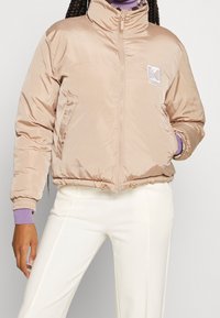 Beżowa kurtka pikowana z wysokim kołnierzem, przednim zamkiem błyskawicznym i bocznymi kieszeniami. Posiada ściągacze na mankietach i wyszywane logo na piersi.