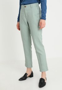 Pantalons en lin vert clair avec une taille haute, jambes droites, poches avant et un bouton marron, associés à des chaussures noires à enfiler.