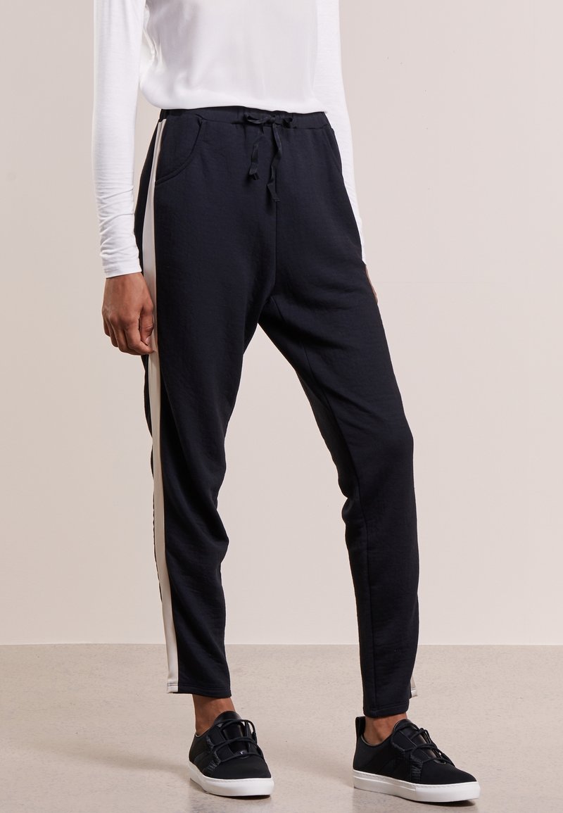 Pantalones jogger negros con rayas blancas laterales, cintura elástica con cordón, usados con zapatillas negras y una camiseta blanca de manga larga.