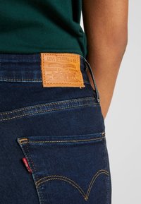 Mörkblå jeans med en brun läderlapp som har präglad text, guldstickning och en röd etikett på bakfickan.