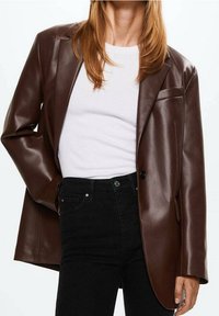 Hnědá faux kožená blazerka s jedním knoflíkem na zapínání, klopovými límci a náprsní kapsou, v kombinaci s bílou košilí a černými džíny.