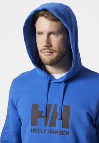 Modrá mikina s kapucí a přední klokanskou kapsou, se velkým černým logem Helly Hansen a nastavitelnou kapucí se šňůrkami.