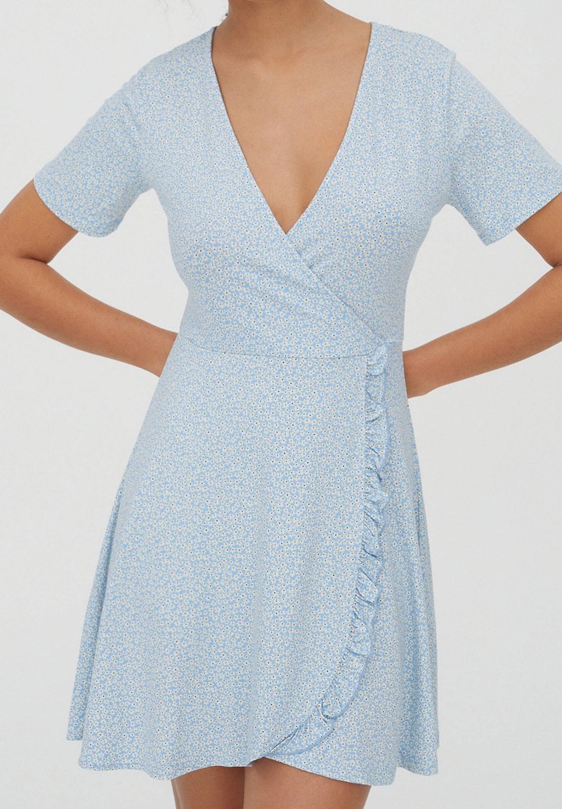 Robe portefeuille bleu clair avec petit motif floral blanc, manches courtes, encolure en V, et détail à volants sur le côté droit devant.