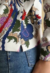 Bluzka w kwiatowy wzór z niebieskimi, czerwonymi i różowymi kwiatami na białym tle, zestawiona z jasnoniebieskimi dżinsami. Teksturowana tkanina, detal z bocznym rozcięciem.