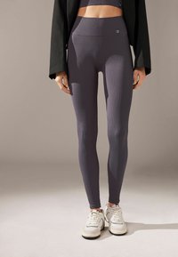 Gerippte lila Leggings mit einem hohen Taillenbund, kombiniert mit einem Cropped-Top und einem schwarzen Blazer, ergänzt durch weiße Sneakers.