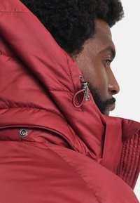Rote Pufferjacke mit hohem Kragen, sichtbaren Nähten, metallischen Beschlägen und verstellbarem Kordelzugdetail an der Kapuze.