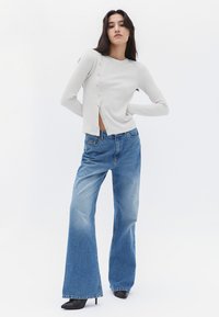 Top a maniche lunghe grigio chiaro a costine con chiusura frontale a bottoni, abbinato a jeans larghi blu sbiaditi e tacchi neri a punta.