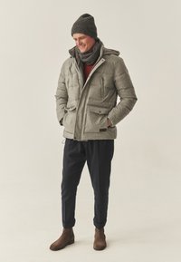 Hall, polsterdatud jakk kõrge kraega, esiküljel tõmblukk ja mitu taskut; kombineeritud tumedate pükste, pruunide saapade ja kootud talvemütsiga.