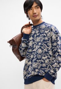 Ναυτικό φούτερ με κρεμ floral σχέδιο, στρογγυλή λαιμόκοψη και ριγέ μανσέτες. Συνδυάζεται με μια καφέ δερμάτινη τσάντα.