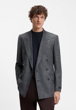 Άνδρας με κατσαρά μαλλιά φοράει γκρι καρό διπλό σακάκι, μαύρο πουκάμισο και σκουρόχρωμο παντελόνι, στέκεται με το ένα χέρι στην τσέπη μπροστά σε λευκό φόντο.
