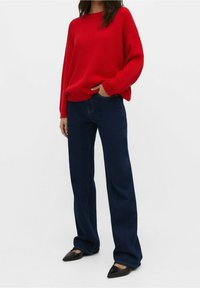 Femme portant un pull rouge côtelé, un jean large bleu foncé et des ballerines noires à bouts pointus, debout avec une main dans la poche.