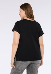 Musta lyhythihainen toppi, jossa on rento istuvuus, pyöreä pääntie ja kontrastiväriset saumat. Yhdistetty harmaisiin farkkuihin.