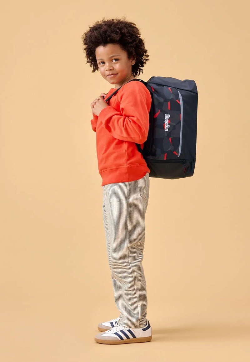 Black backpack with red accents, fabric material, featuring padded straps. The child wears an orange sweatshirt and striped pants, standing sideways.